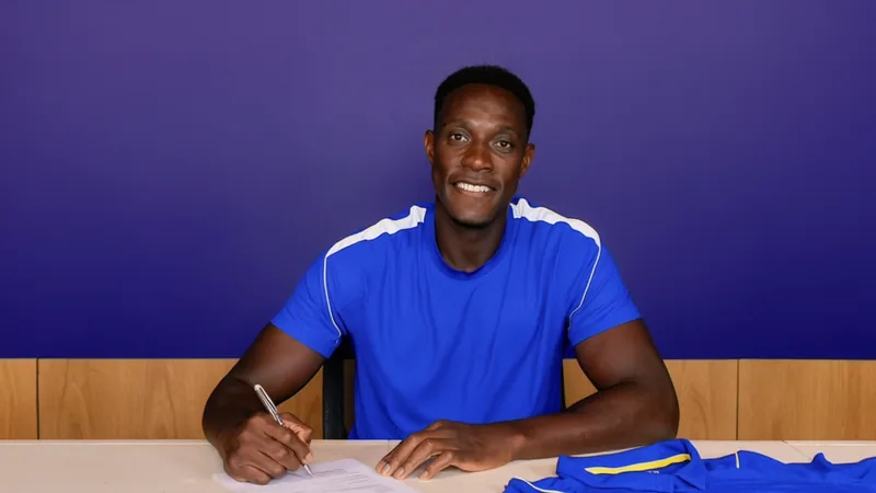 Danny Welbeck sorrindo com a camisa do Chelsea em frente ao estádio Stamford Bridge, com o gramado ao fundo e a torcida animada nas arquibancadas.
