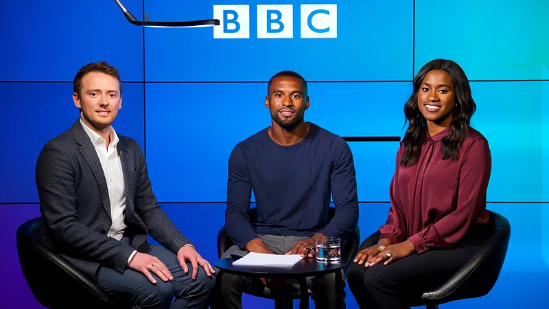 Fraizer Campbell no Lie Detector da BBC — Football World Cup News Fraizer Campbell sorrindo em um estúdio de podcast da BBC, com um detector de mentiras e um microfone à sua frente, durante a gravação do Lie