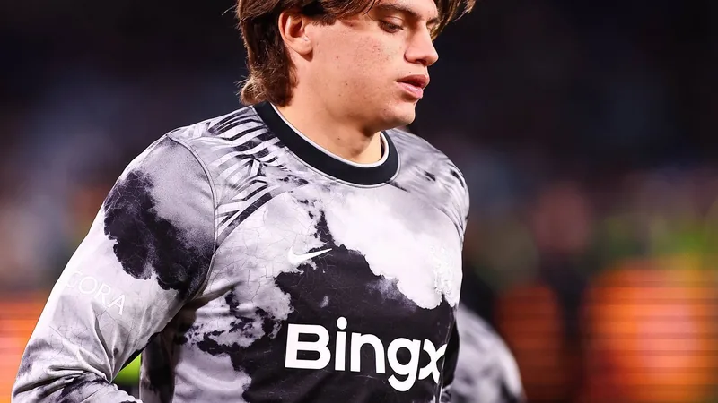 Jovem jogador Marco Palestra vestindo a camisa do Chelsea, sorrindo em frente ao estádio Stamford Bridge, com o técnico Xabi Alonso ao fundo.