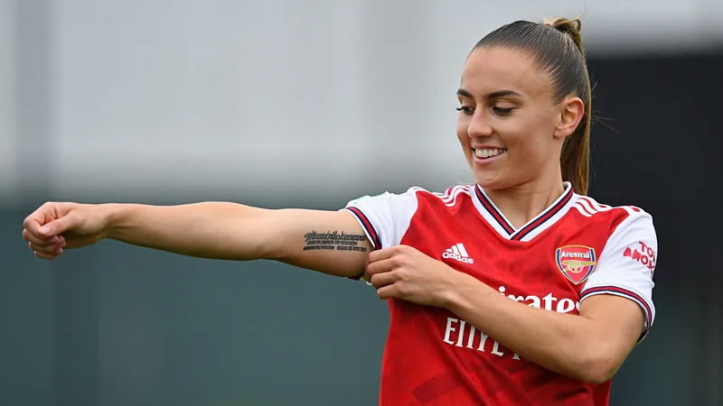 Georgia Stanway tatua hino do Arsenal após chegar ao clube — Football Georgia Stanway sorri mostrando a tatuagem 'Whatever the weather' no braço direito, com a camisa do Arsenal ao fundo no Emirates Stadium.