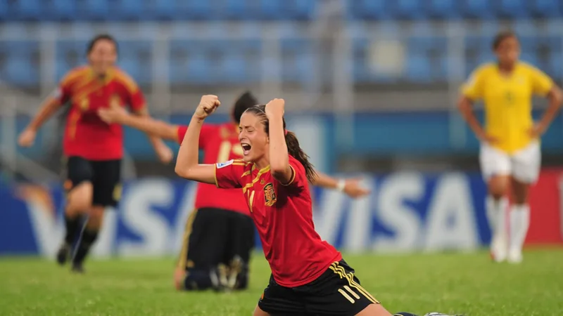 Jogadoras de futebol feminino sub-17 em ação durante uma partida do Mundial, com estádio lotado e bandeiras dos países ao fundo.